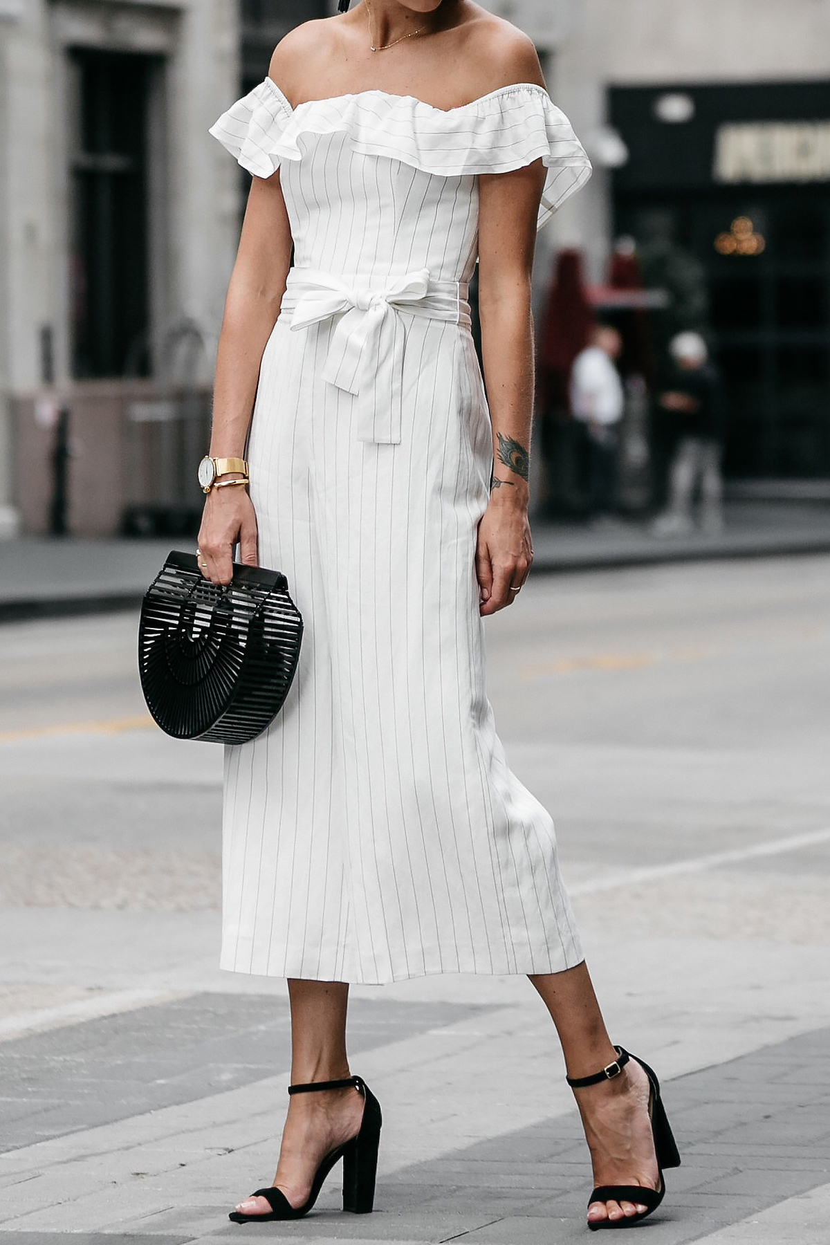 Blonde woman wearing Club Monaco off-the-shoulder ruffle white jumpsuit Cult Gaia Black Acrylic Ark Clutch Black Ankle Strap Heeled Sandals